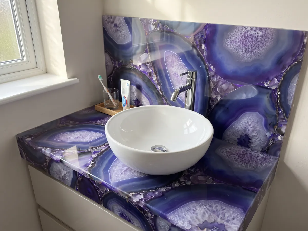 Cubierta de lavabo y pared de baño en Ágata Morada con lavamanos blanco y grifería cromada: rodajas grandes de ágata violeta azulado con centros lila cristalinos
