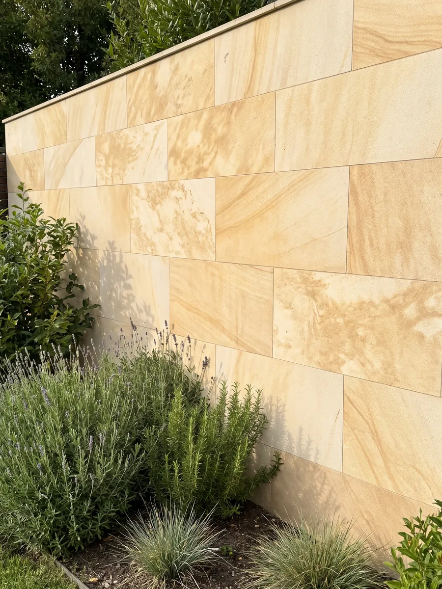Muro de jardín revestido con Arenisca Santafiora Havana con plantas delante: tono crema cálido con nubes doradas difusas y textura arenosa suave