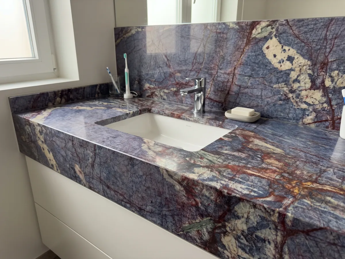 Cubierta de lavabo y pared de baño en cuarcita azul Blue Luise con lavamanos blanco y grifería cromada: azul violáceo atravesado por una red densa de vetas rojo óxido