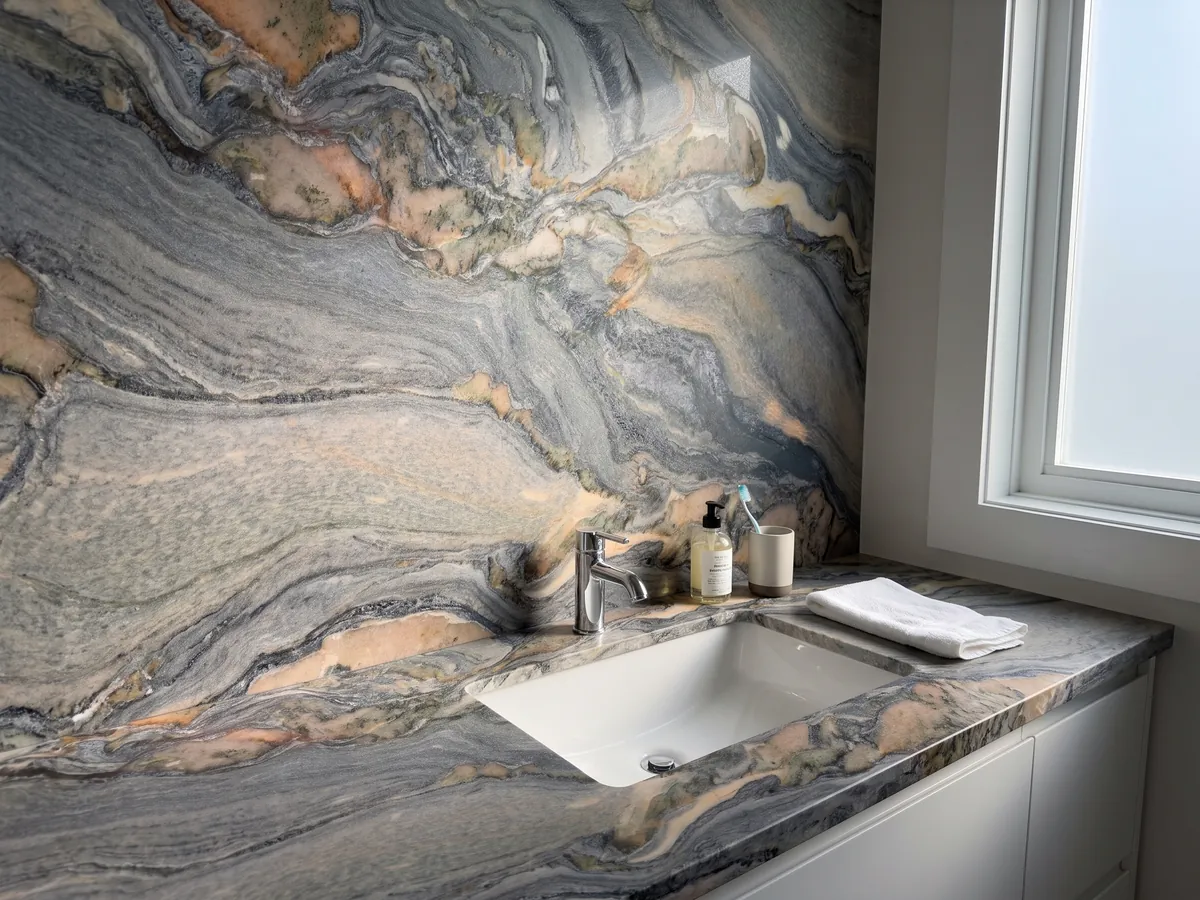 Cubierta de lavabo y pared de baño en cuarcita azul Caravaggio con lavamanos blanco y grifería cromada: remolinos gris azulados con manchas salmón y crema en movimiento diagonal