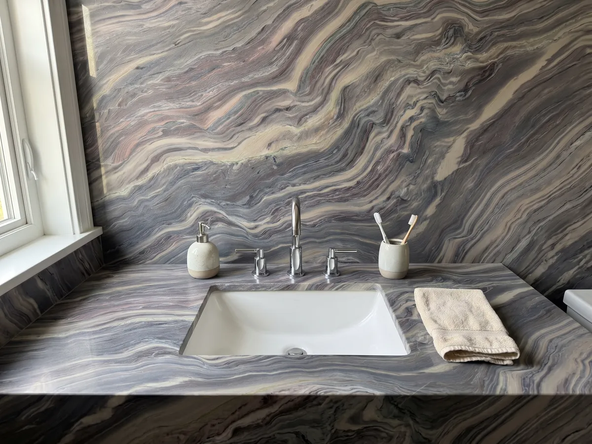 Cubierta de lavabo y pared de baño en cuarcita azul Explosion Blue con lavamanos blanco y grifería cromada: ondas apretadas gris azuladas y crema que recorren toda la placa en diagonal