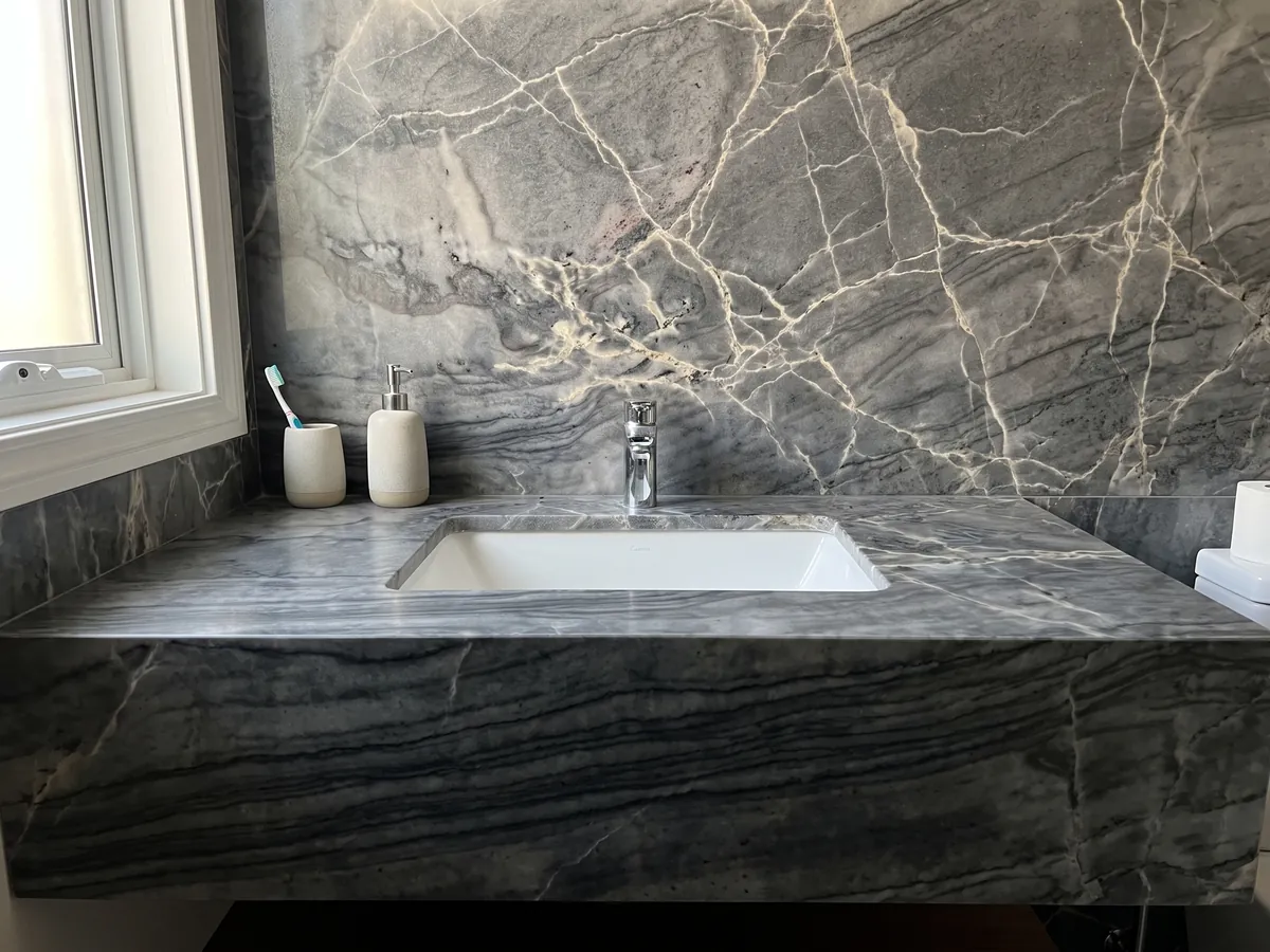 Cubierta de lavabo y pared de baño en cuarcita gris London Grey con lavamanos blanco y grifería cromada: gris carbón con red de vetas crema y ondas horizontales oscuras
