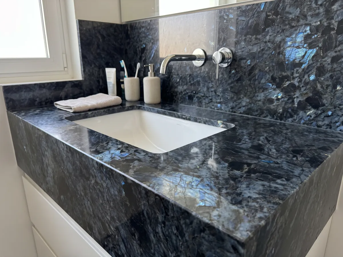 Cubierta de lavabo y pared de baño en cuarcita negra Lemurian con lavamanos blanco y grifería cromada: negro de cristales grandes con destellos azul iridiscente