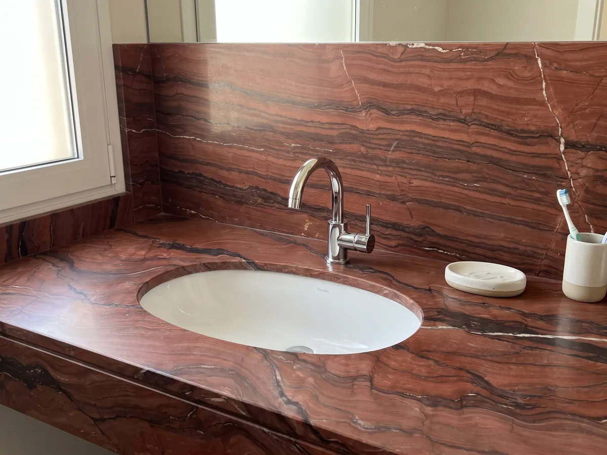 Cubierta de lavabo y pared de baño en cuarcita roja Fusion Red con lavamanos blanco y grifería cromada: rojo ladrillo con ondas oscuras horizontales y una fina veta blanca