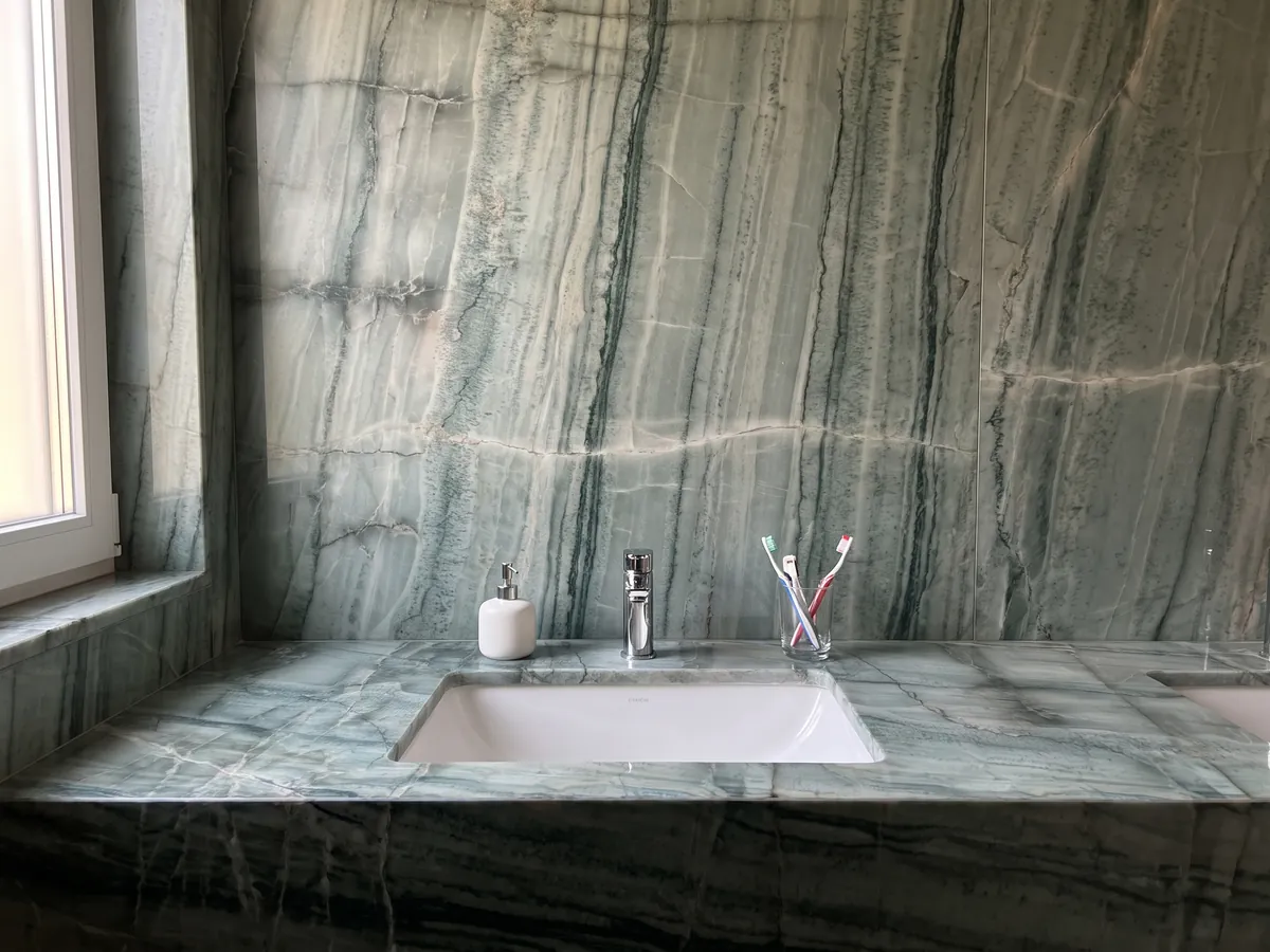 Cubierta de lavabo y pared de baño en cuarcita verde da Vinci con lavamanos blanco y grifería cromada: verde grisáceo con cuadrícula de vetas oscuras verticales y bandas claras