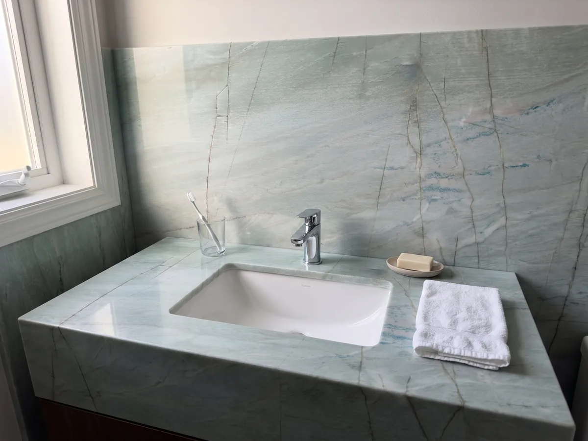 Cubierta de lavabo y pared de baño en cuarcita verde Emerald Green con lavamanos blanco y grifería cromada: verde agua claro con finas líneas oscuras verticales y pequeños destellos azules