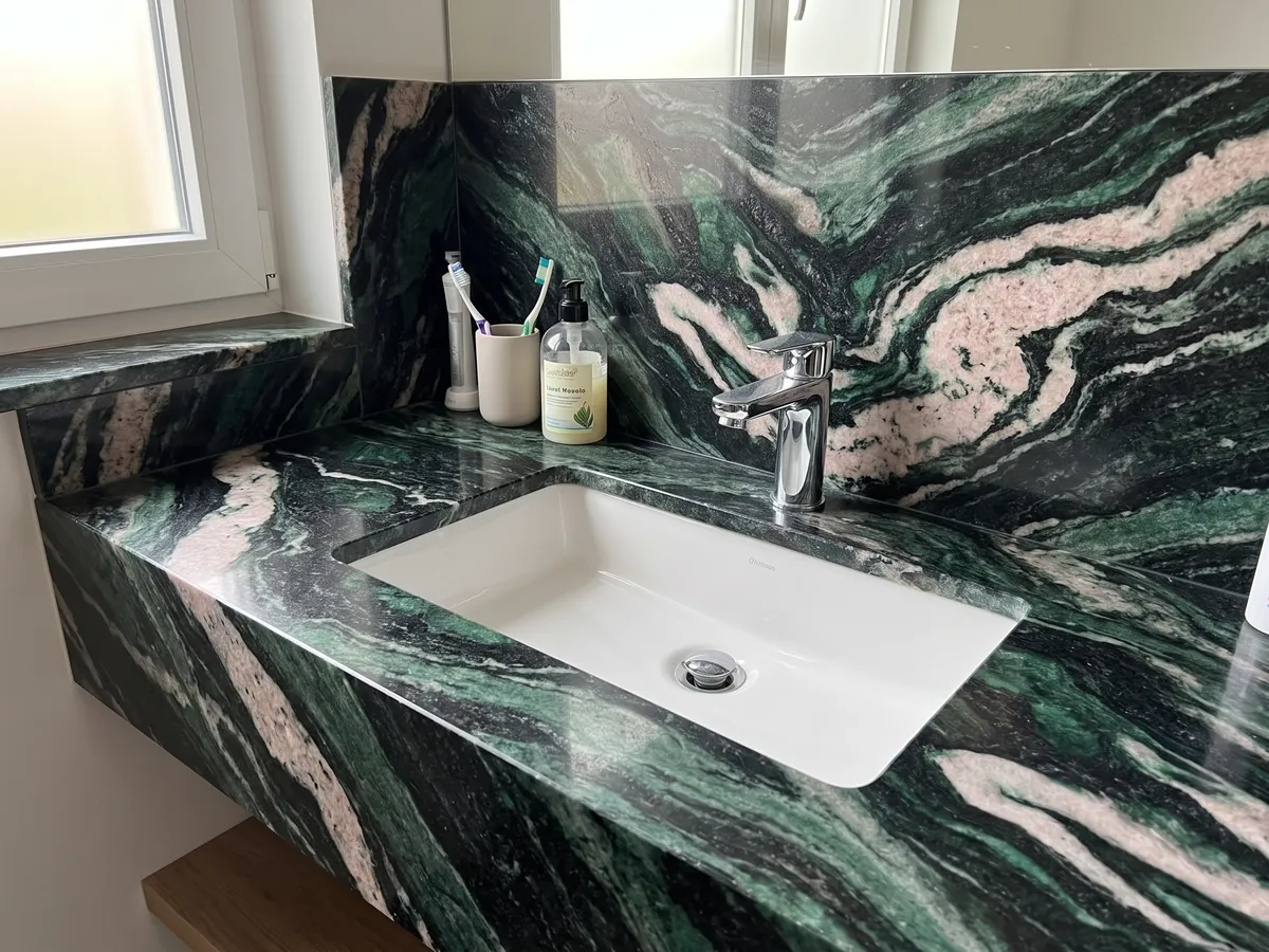 Cubierta de lavabo y pared de baño en cuarcita verde Lapponia Green con lavamanos blanco y grifería cromada: verde esmeralda y negro con grandes ondas blanco rosadas