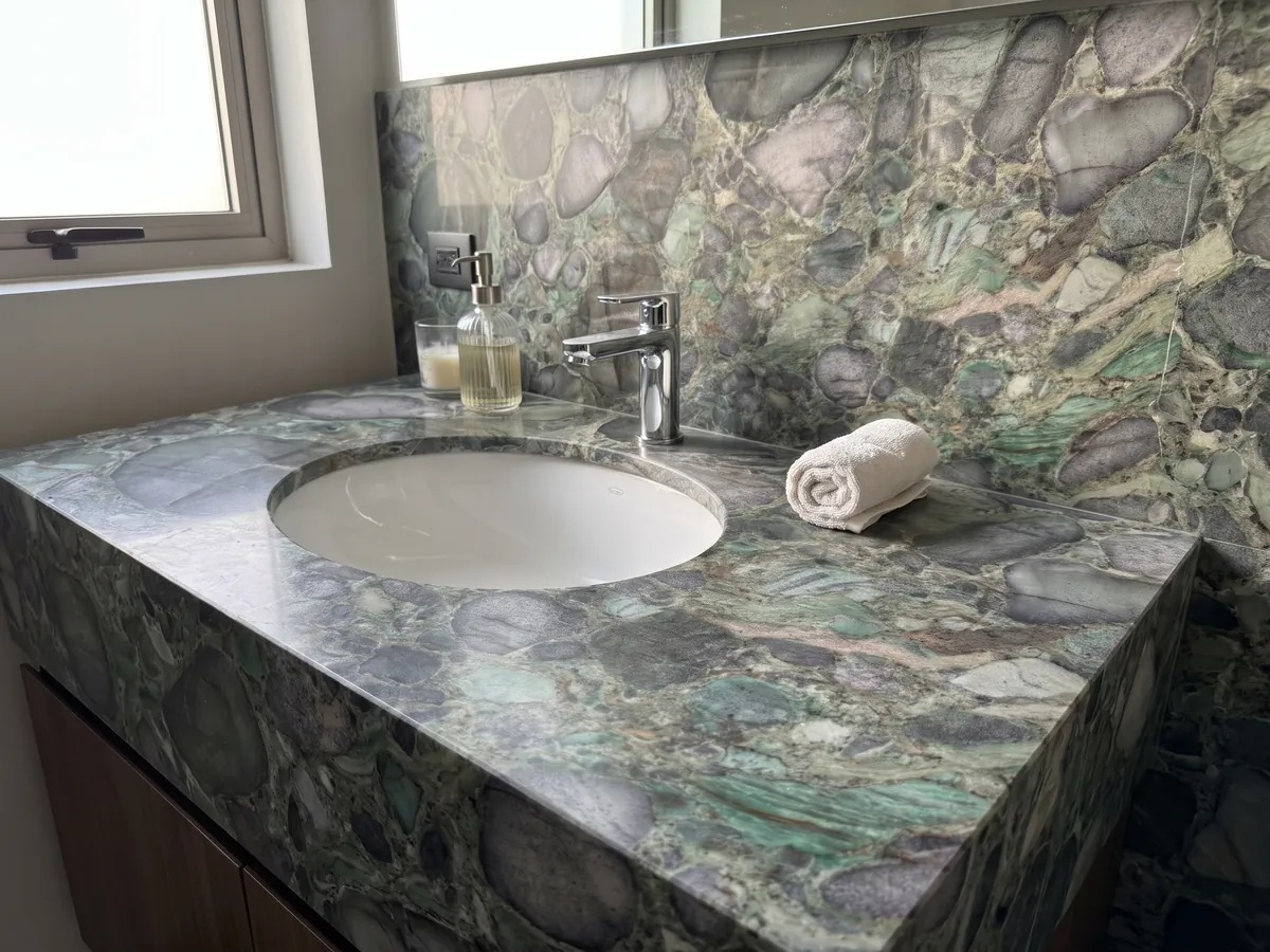 Cubierta de lavabo y pared de baño en cuarcita verde Palladium Green con lavamanos blanco y grifería cromada: clastos redondeados gris violáceo con matriz verde y destellos esmeralda