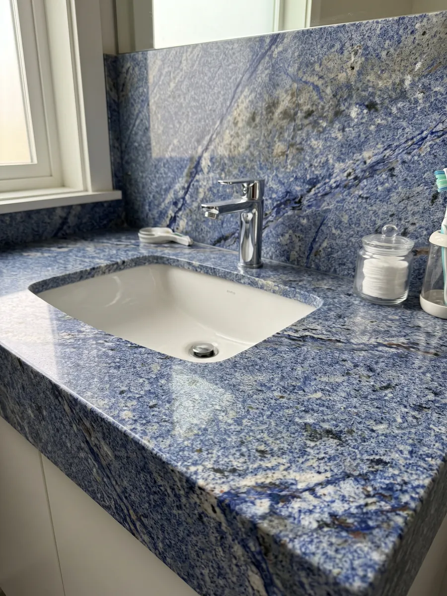 Cubierta de lavabo y pared de baño en Granito Azul Bahia con lavamanos blanco y grifería cromada: azul intenso granulado con zonas blancas y manchas oscuras