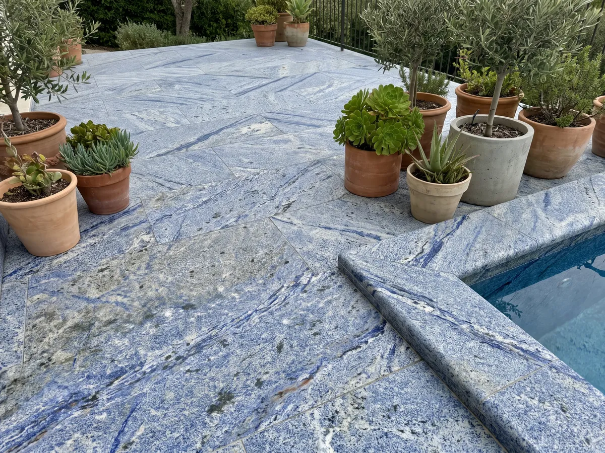 Piso de terraza y borde de piscina en Granito Azul Bahia con macetas: azul intenso granulado con zonas blancas y manchas oscuras