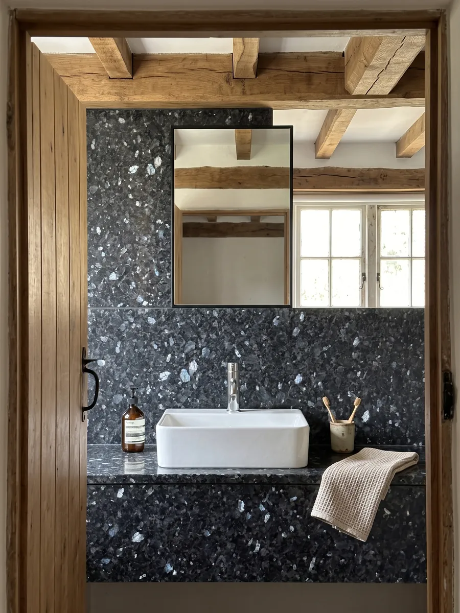 Cubierta de lavabo y pared de baño en granito azul Blue Pearl con lavamanos blanco y grifería cromada: gris azulado casi negro con cristales que brillan azul plateado