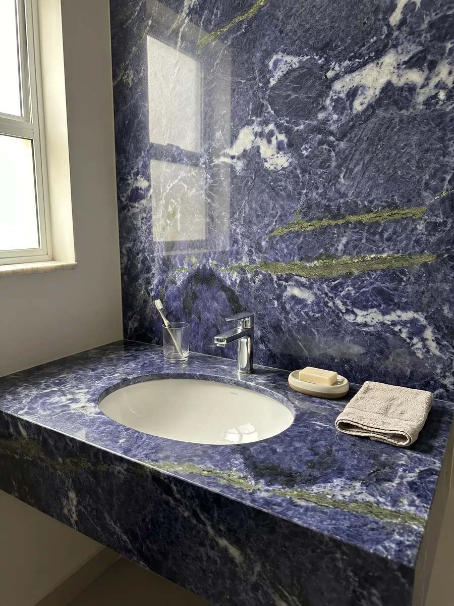 Cubierta de lavabo y pared de baño en granito azul Jeriba Blue con lavamanos blanco y grifería cromada: azul violeta intenso con nubes blancas y una línea verde dorada