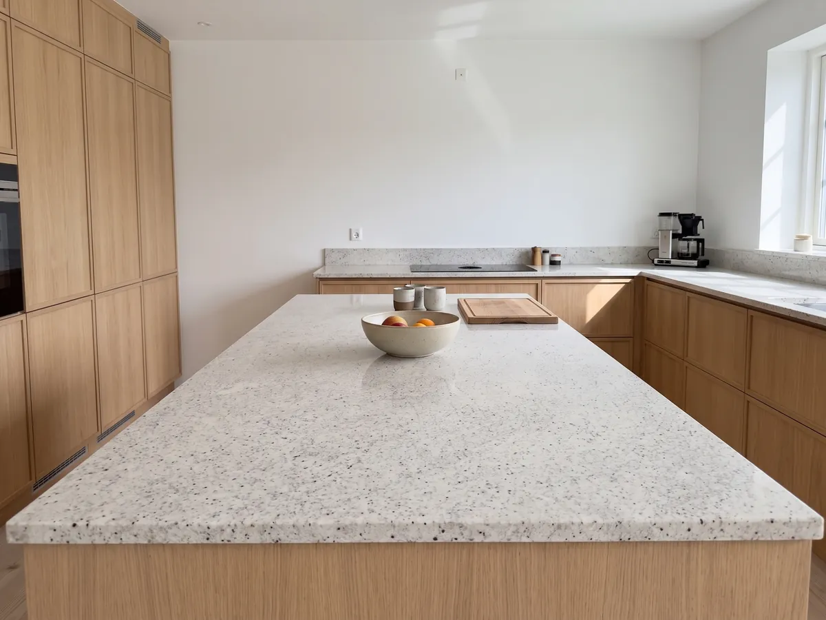 Isla de cocina con cubierta de Granito Blanco Dallas y canto recto en una cocina con luz natural: blanco cremoso con un moteado fino gris y pequeños puntos burdeos