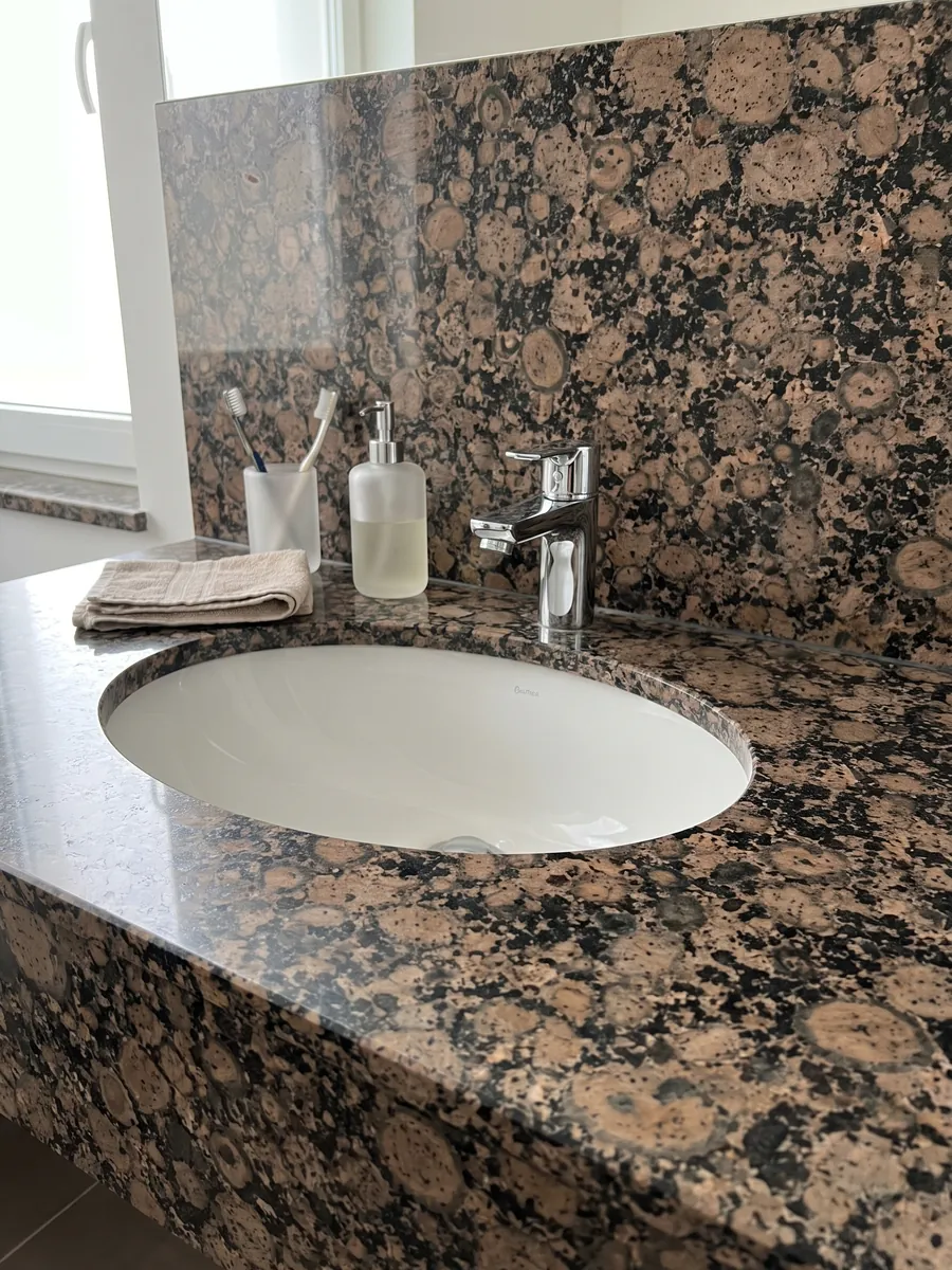 Cubierta de lavabo y pared de baño en granito café Baltic Brown con lavamanos blanco y grifería cromada: granos redondos marrones con anillos sobre fondo moteado negro