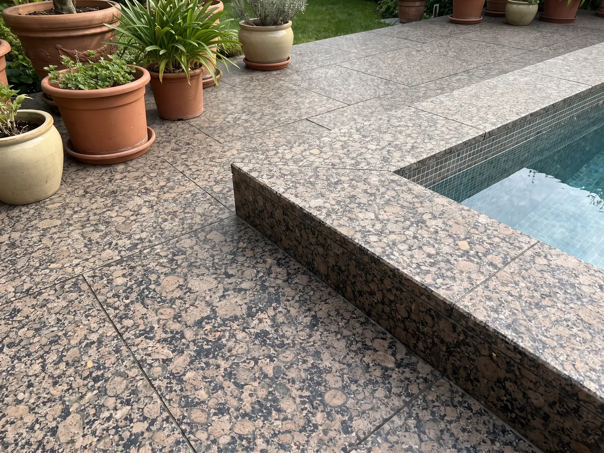 Piso de terraza y borde de piscina en granito café Baltic Brown con macetas: granos redondos marrones con anillos sobre fondo moteado negro