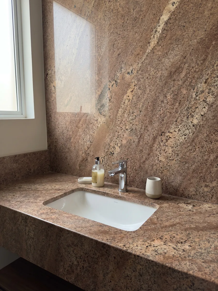 Cubierta de lavabo y pared de baño en granito café Juparana Florence con lavamanos blanco y grifería cromada: marrón cobrizo granulado con zonas beige difusas