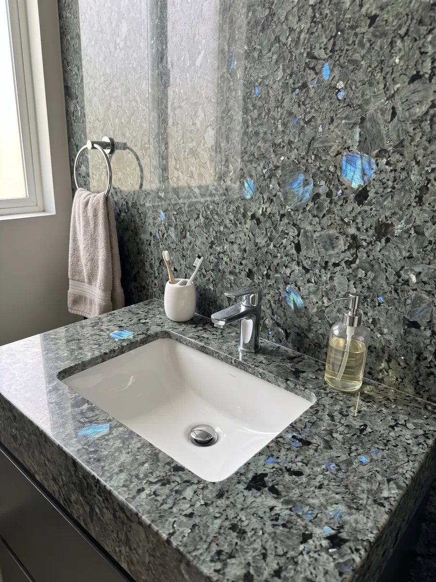 Cubierta de lavabo y pared de baño en granito gris Labradorita con lavamanos blanco y grifería cromada: cristales grandes gris verdosos con destellos azules iridiscentes