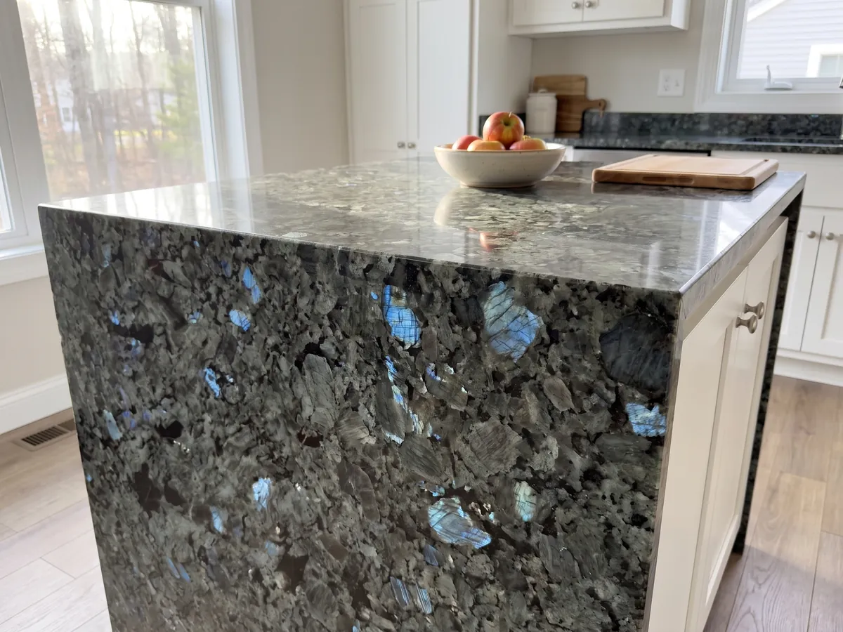 Isla de cocina con cubierta de granito gris Labradorita y canto recto en una cocina con luz natural: cristales grandes gris verdosos con destellos azules iridiscentes