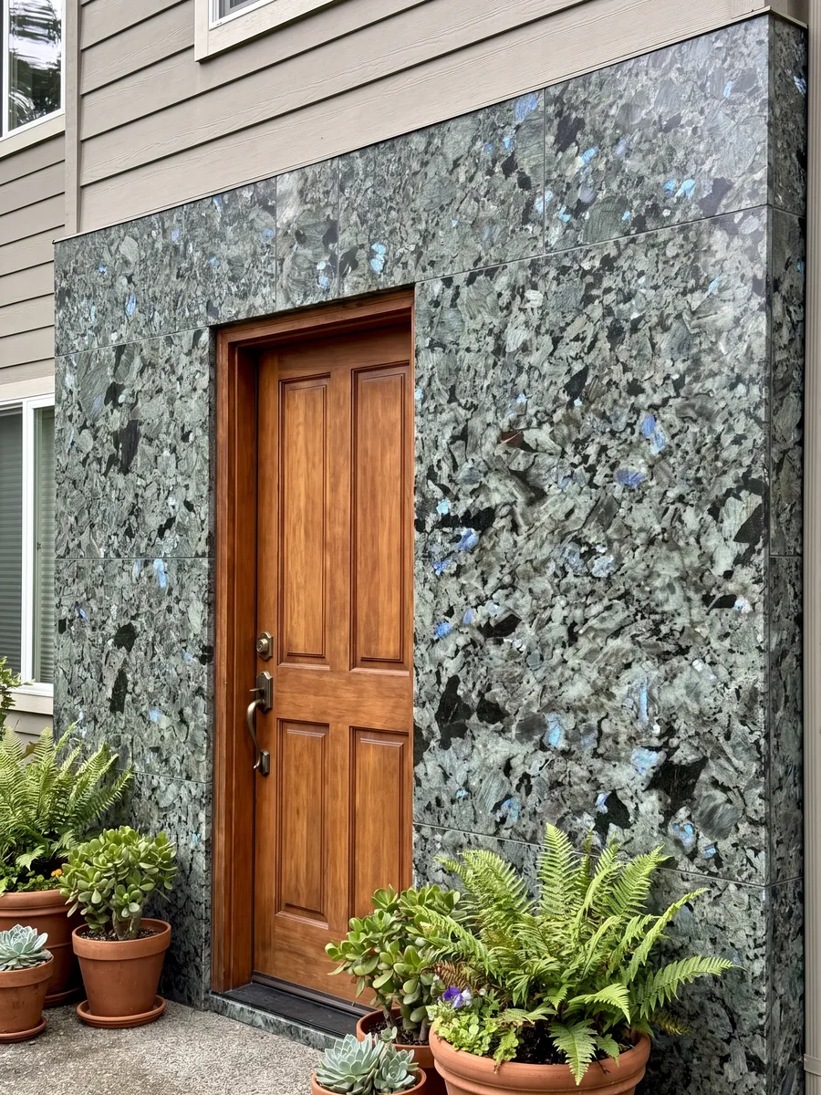 Fachada de casa revestida con granito gris Labradorita junto a una puerta de madera: cristales grandes gris verdosos con destellos azules iridiscentes