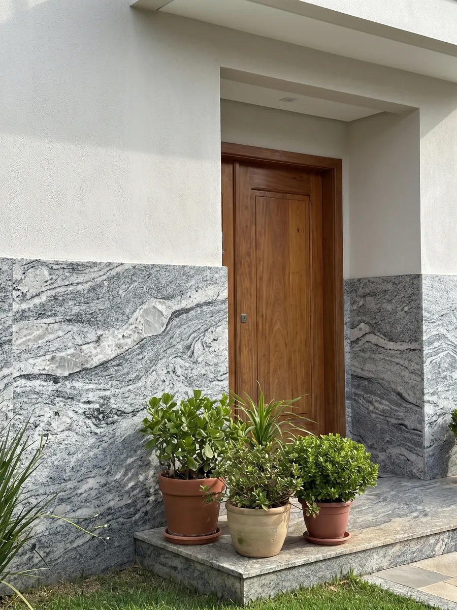 Fachada de casa revestida con granito gris Piracema White junto a una puerta de madera: bandas onduladas gris y blanco con un gran fragmento claro