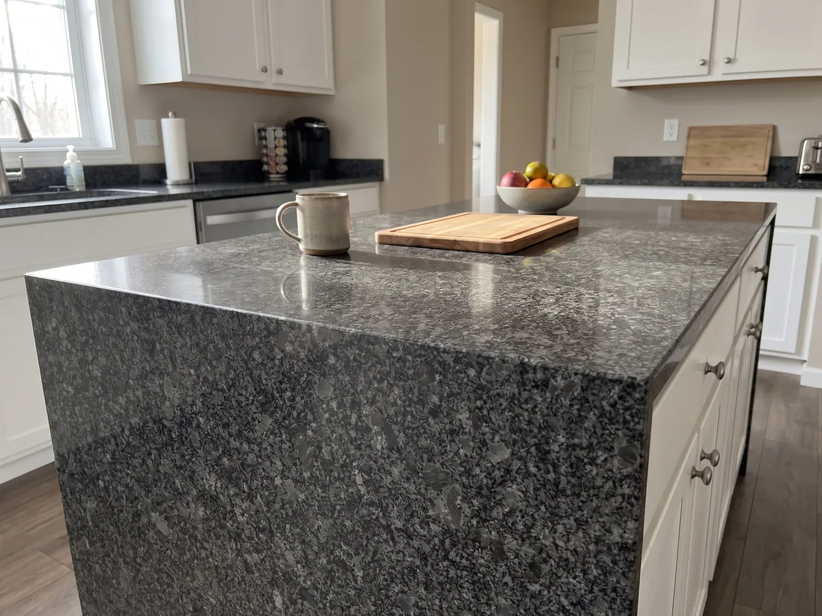 Isla de cocina con cubierta de granito gris Steel Grey y canto recto en una cocina con luz natural: grano medio gris acero sobre fondo casi negro, con pequeñas áreas grises más claras repartidas