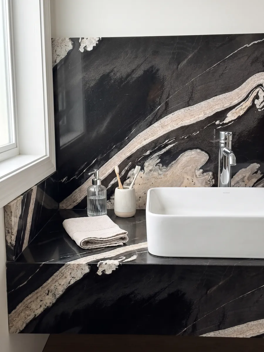 Cubierta de lavabo y pared de baño en granito negro Copacabana con lavamanos blanco y grifería cromada: negro con una banda crema ondulada que cruza en diagonal