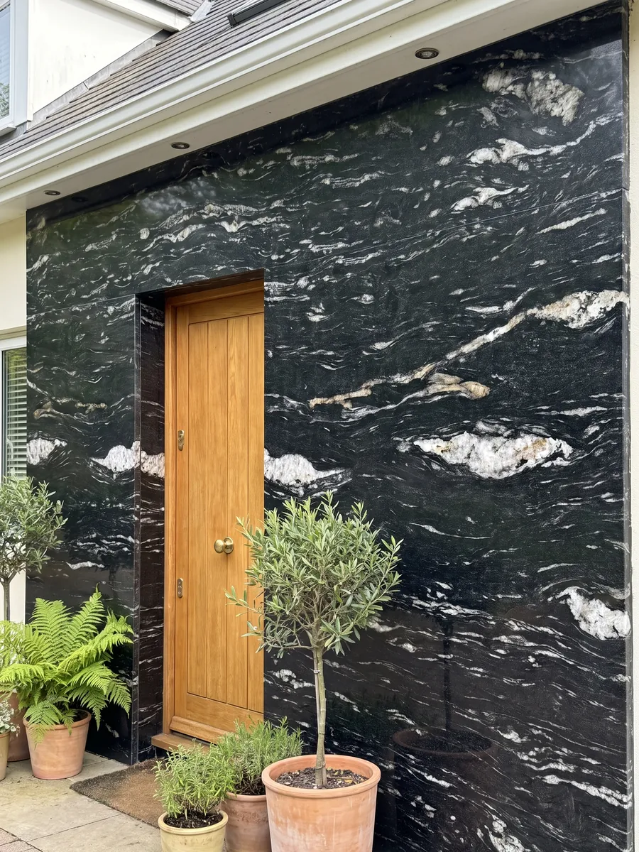 Fachada de casa revestida con granito negro Cosmic Black junto a una puerta de madera: negro con trazos blancos ondulados horizontales