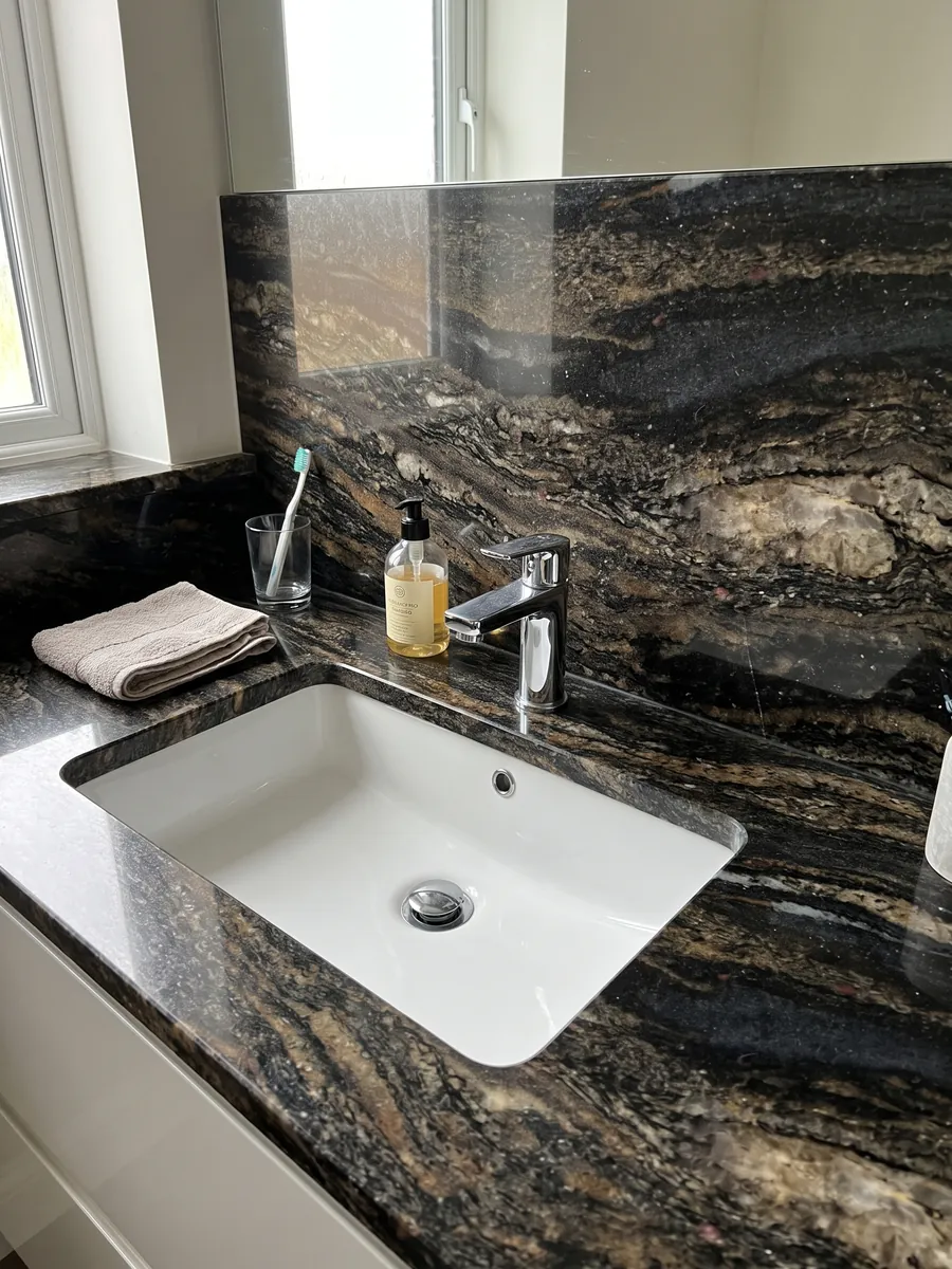 Cubierta de lavabo y pared de baño en granito negro Cosmic Gold con lavamanos blanco y grifería cromada: negro con zonas doradas y bronce en ondas horizontales