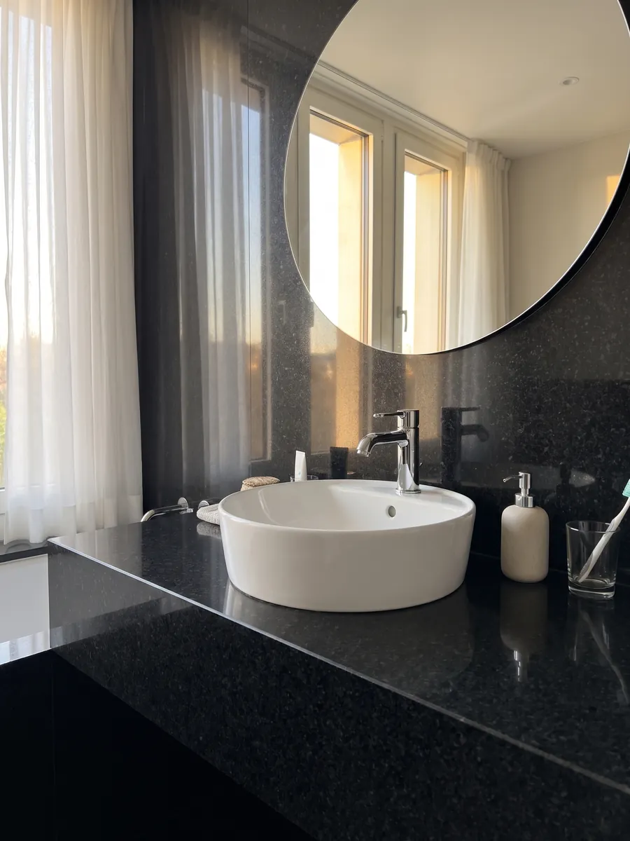 Cubierta de lavabo y pared de baño en Granito Negro San Gabriel con lavamanos blanco y grifería cromada: negro profundo de grano medio con motas gris oscuro que se ven de cerca