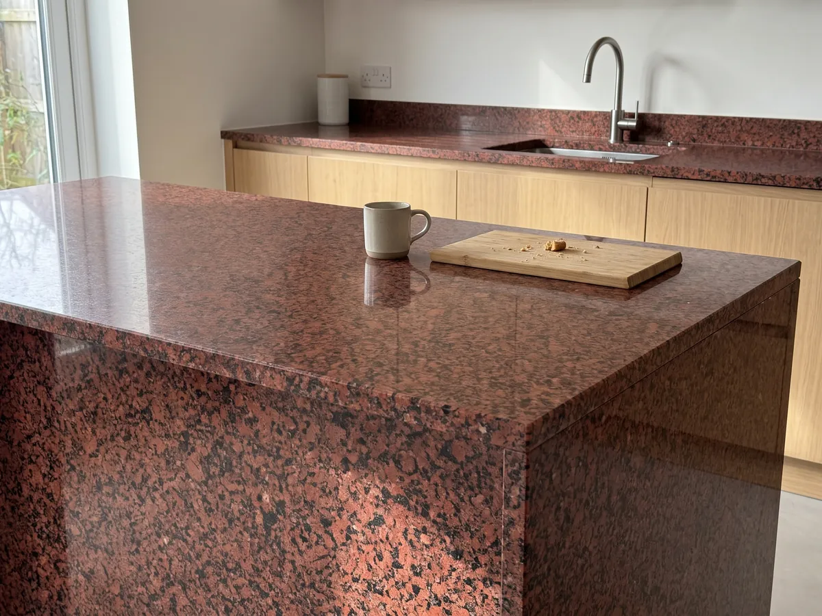 Isla de cocina con cubierta de granito rojo Balmoral Red y canto recto en una cocina con luz natural: rojo intenso granulado con puntos negros repartidos