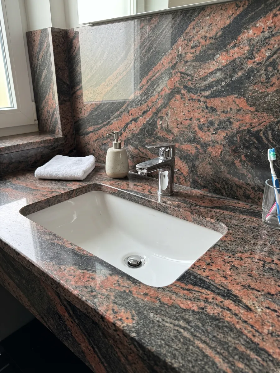 Cubierta de lavabo y pared de baño en granito rojo Indian Aurora con lavamanos blanco y grifería cromada: ondas de color coral y gris oscuro en diagonal