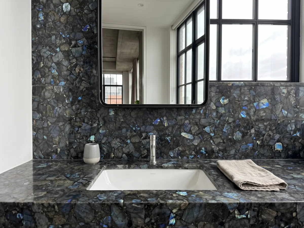Cubierta de lavabo y pared de baño en Labradorita con lavamanos blanco y grifería cromada: piezas gris oscuro con destellos azules iridiscentes repartidos