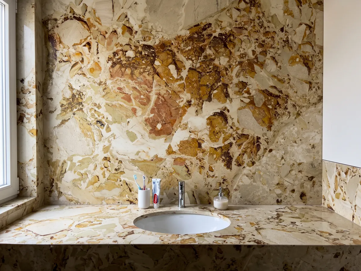 Cubierta de lavabo y pared de baño en mármol beige Alma Fuoco con lavamanos blanco y grifería cromada: manchas ocre y óxido de bordes recortados que se agrupan en la parte superior central