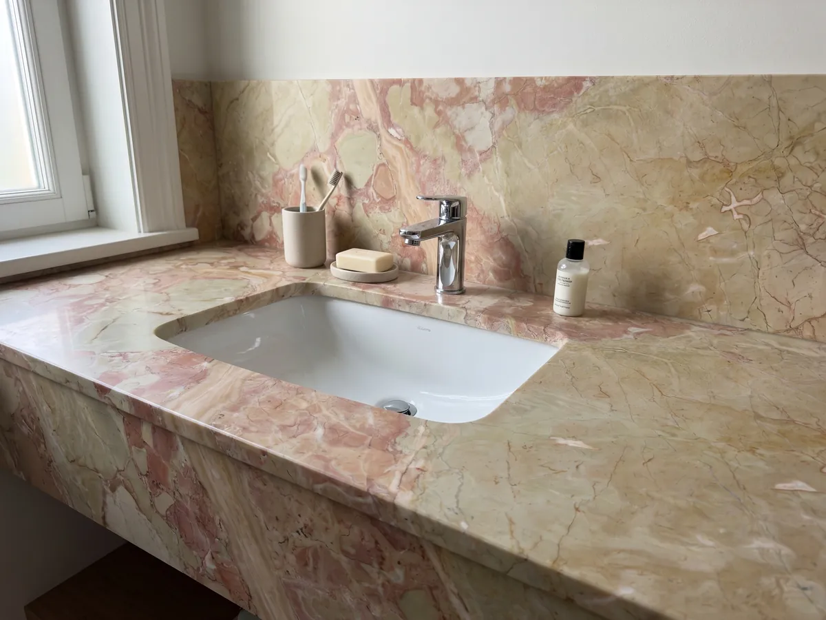 Cubierta de lavabo y pared de baño en mármol beige Breccia D’Aragona con lavamanos blanco y grifería cromada: fragmentos beige verdosos grandes con manchas rosa coral en el borde izquierdo