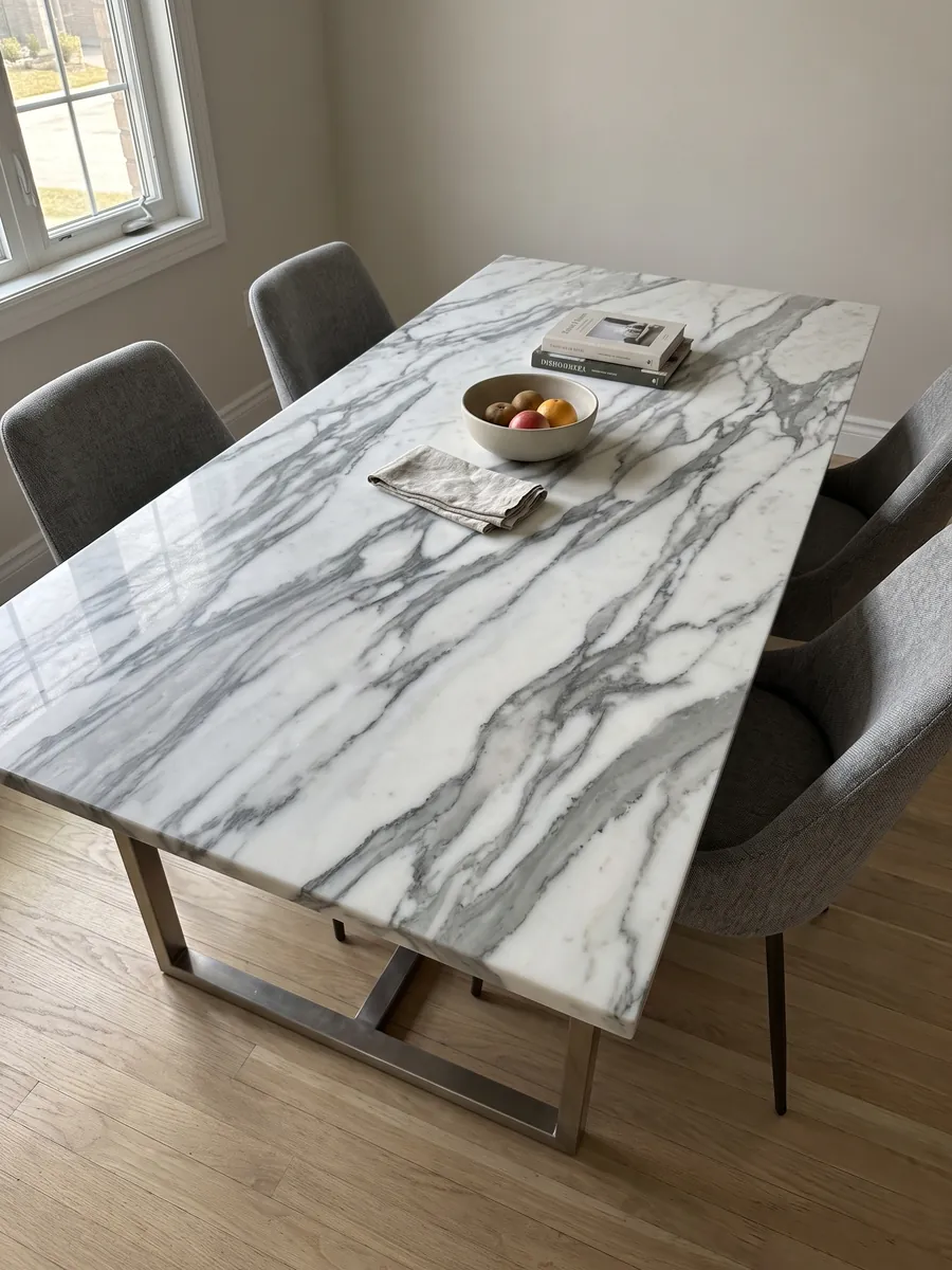 Mesa de comedor con cubierta de mármol blanco Arabescato Apuano sobre base de acero: vetas gris oscuro gruesas y ramificadas que cruzan el blanco en diagonal