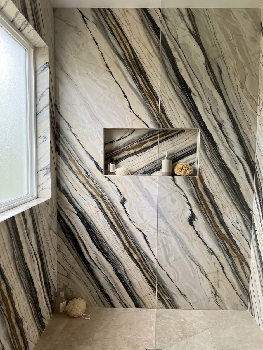 Ducha revestida con placas enteras de mármol blanco Calacatta Oyster con nicho empotrado: bandas anchas negras y marrones paralelas en diagonal sobre crema