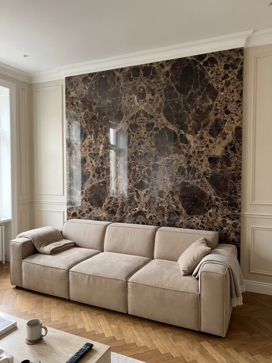 Pared de sala revestida con placas de mármol café Emperador Oscuro abiertas en libro detrás de un sofá: manchas marrón oscuro casi negras unidas por una red densa beige