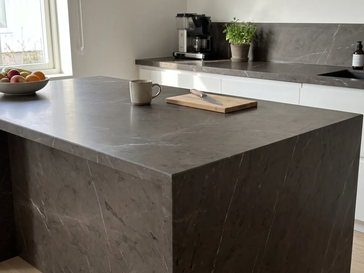 Isla de cocina con cubierta de mármol café Grafite y canto recto en una cocina con luz natural: fondo oscuro entre marrón y grafito con pocas vetas blancas muy finas