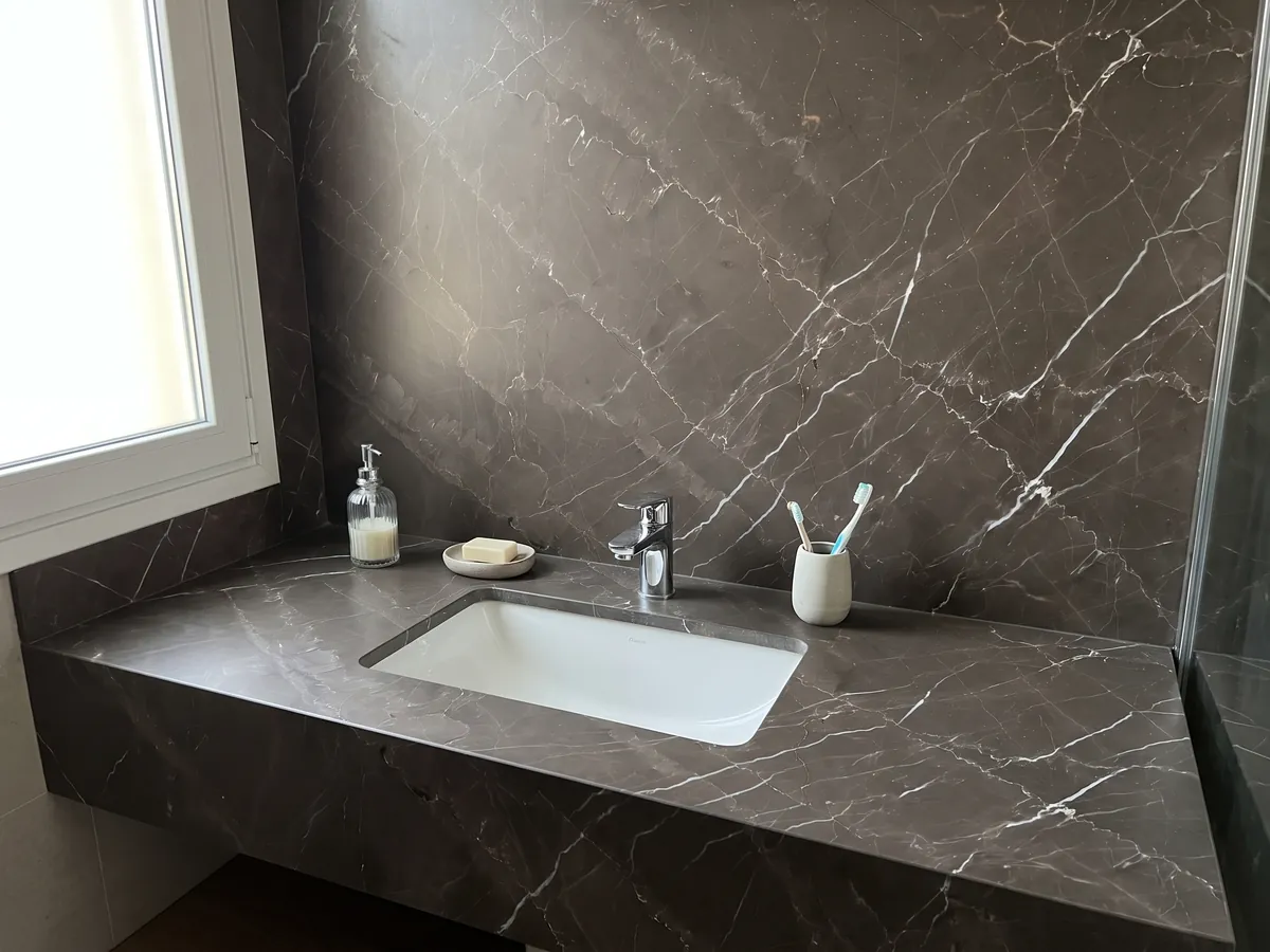 Cubierta de lavabo y pared de baño en Mármol Gris Amani con lavamanos blanco y grifería cromada: fondo topo oscuro cruzado por vetas blancas finas en diagonal