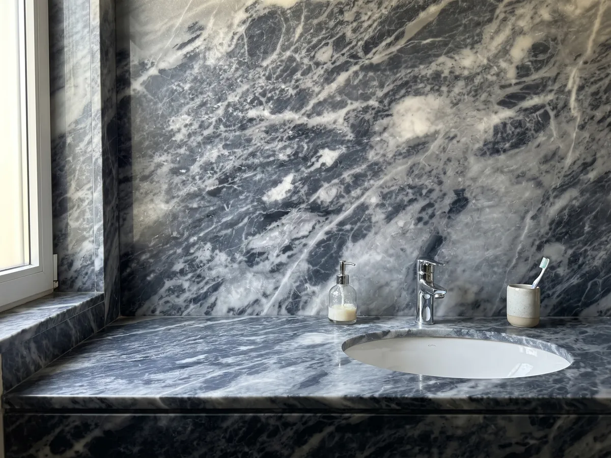 Cubierta de lavabo y pared de baño en mármol gris Bardiglio Nuvolato con lavamanos blanco y grifería cromada: gris oscuro azulado con nubes y vetas blanquecinas difusas en diagonal