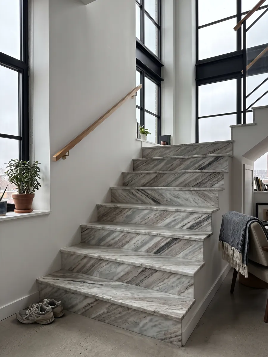 Escalera interior con peldaños de mármol gris Fantasy Brown con pasamanos: nubes alargadas de blanco y gris con algunos trazos oscuros en diagonal