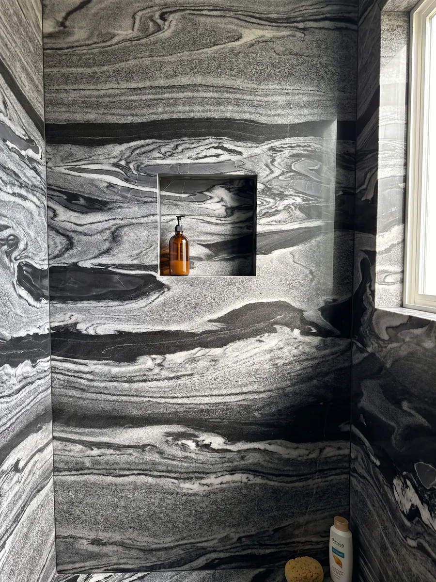 Ducha revestida con placas enteras de Mármol Gris Imperial con nicho empotrado: ondas horizontales grises y blancas sobre negro, como corrientes en movimiento