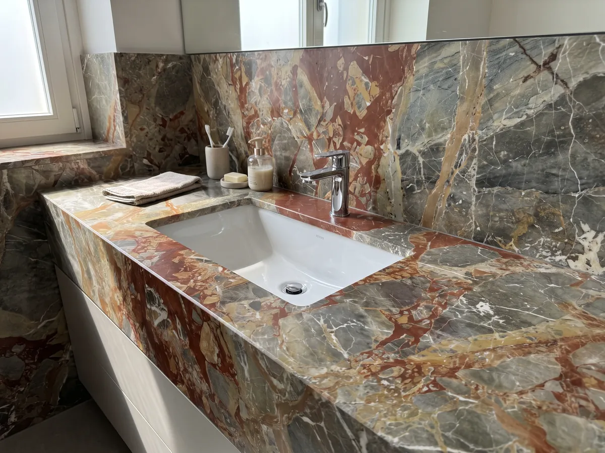 Cubierta de lavabo y pared de baño en mármol gris Macchia Vecchia con lavamanos blanco y grifería cromada: fragmentos gris verdoso con grandes manchas rojo óxido y vetas blancas