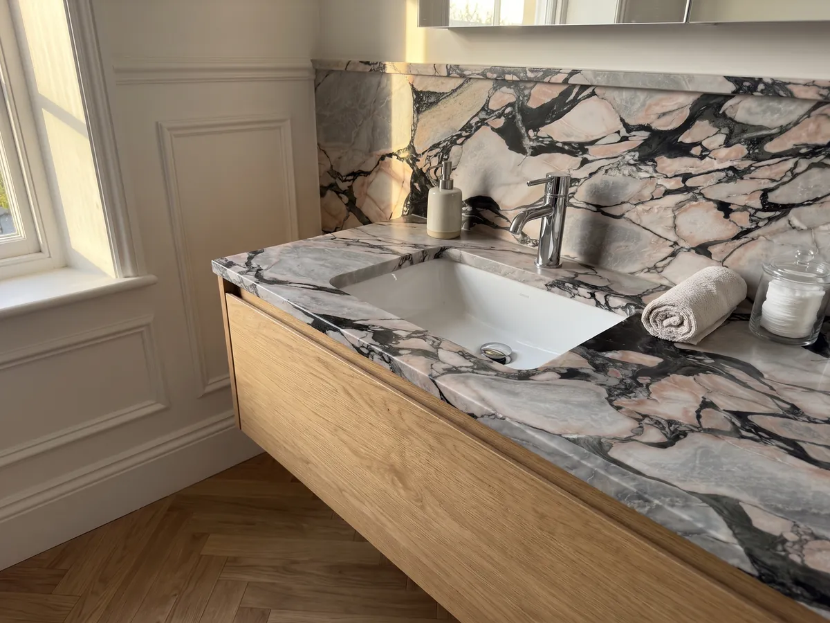 Cubierta de lavabo y pared de baño en mármol gris Notre Dame con lavamanos blanco y grifería cromada: fragmentos grises y rosa salmón encerrados por vetas negras gruesas