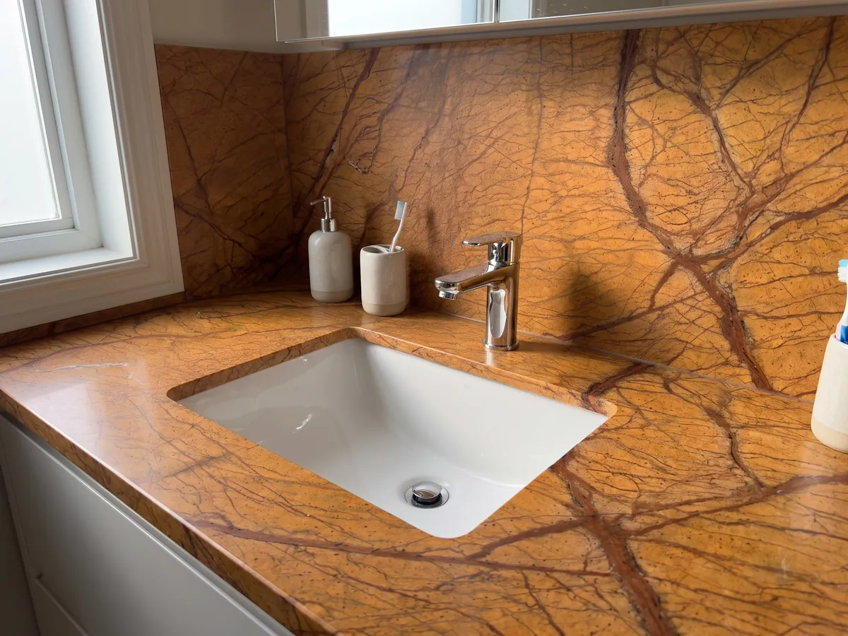 Cubierta de lavabo y pared de baño en mármol naranja Picasso Gold con lavamanos blanco y grifería cromada: naranja ocre intenso con una red de vetas marrón rojizo ramificadas