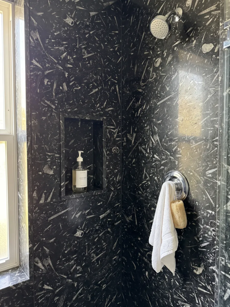 Ducha revestida con placas enteras de mármol negro Fossil Black con nicho empotrado: fondo negro lleno de fósiles alargados en forma de cono, de color gris claro