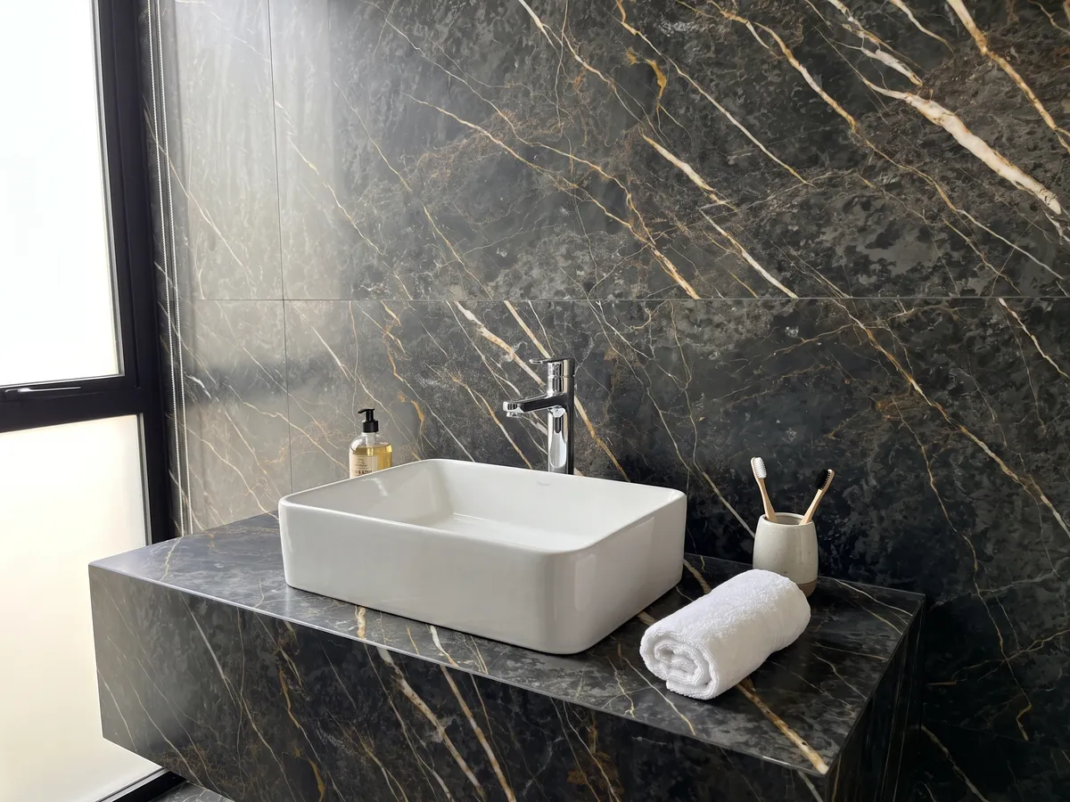 Cubierta de lavabo y pared de baño en mármol negro Port Laurent con lavamanos blanco y grifería cromada: gris carbón con vetas doradas y blancas paralelas en diagonal
