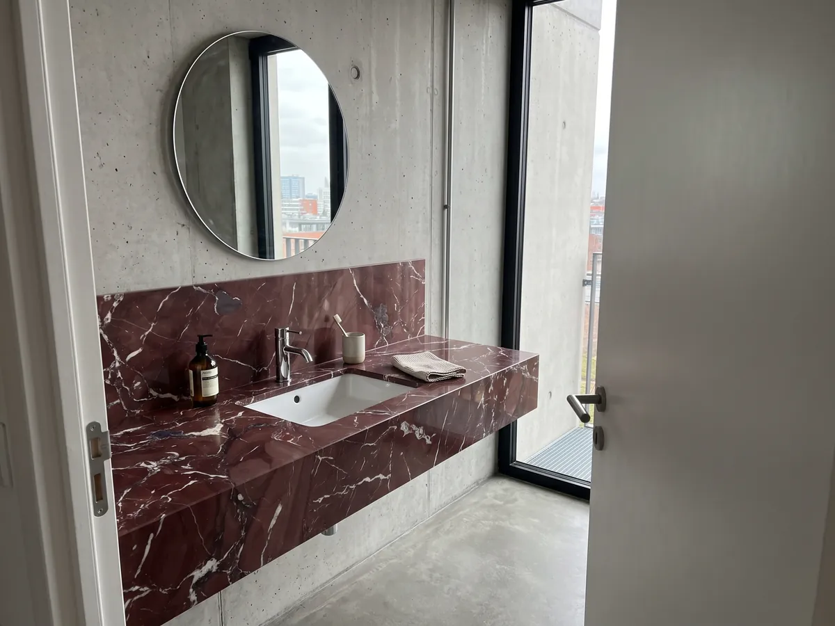 Cubierta de lavabo y pared de baño en Mármol Rojo Cardinale con lavamanos blanco y grifería cromada: rojo burdeos con vetas blancas irregulares y algunas manchas gris violáceo