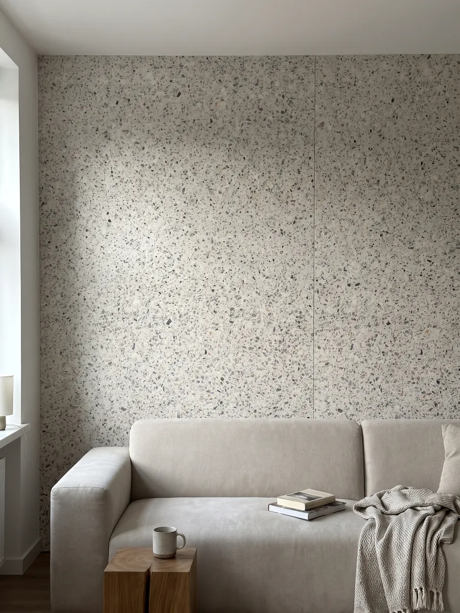Pared de sala revestida con placas de Mármol Sintético Fior di Pesco White con Resina abiertas en libro detrás de un sofá: fondo marfil con fragmentos grises y oscuros repartidos, estilo terrazo