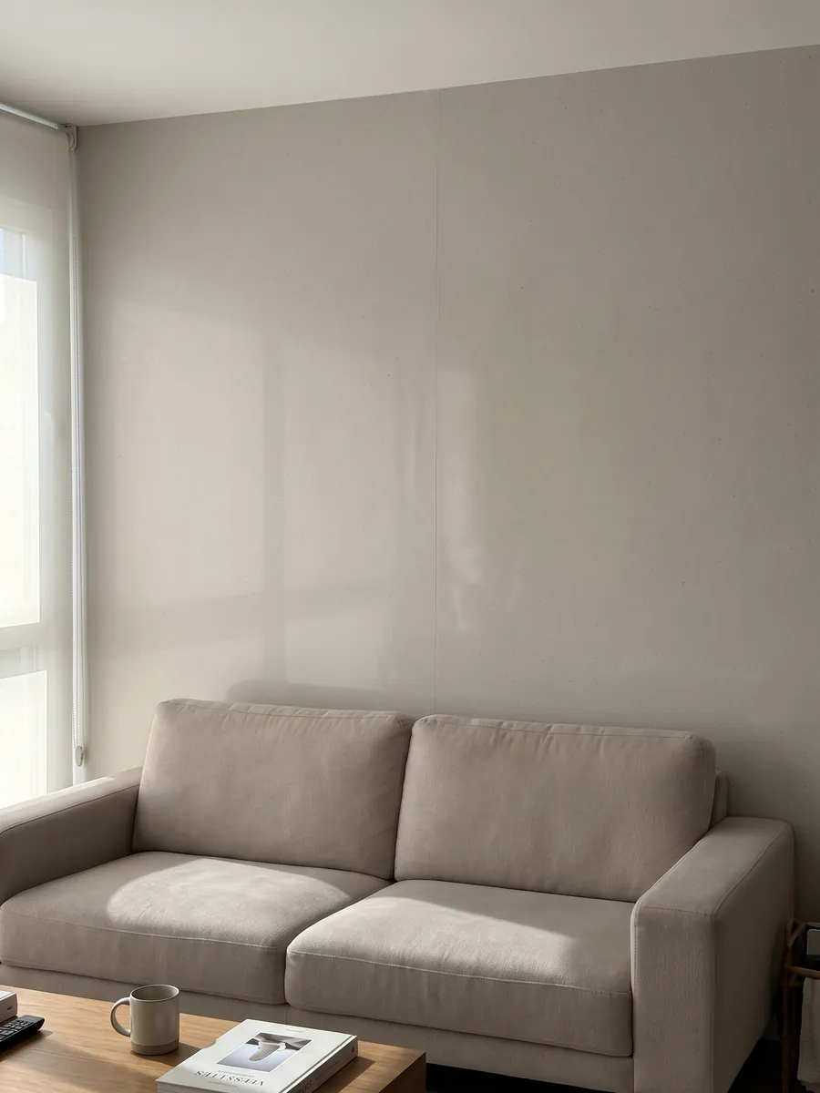 Pared de sala revestida con placas de Mármol Sintético Veselje con Resina abiertas en libro detrás de un sofá: color liso gris perla cálido; la imagen no muestra textura, solo un degradado de luz